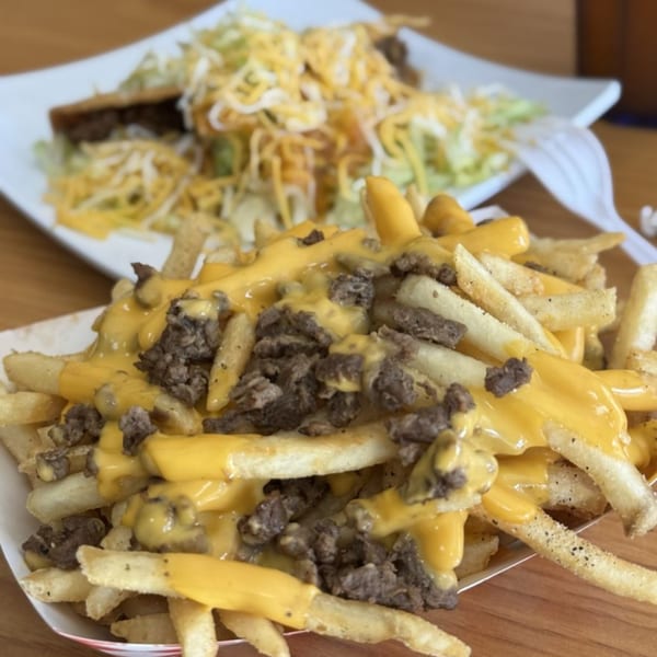 Cheesy fries topped with steak, with a plate of tacos in the blurred background.
