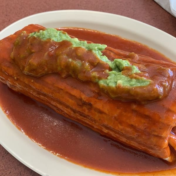 A large, saucy Mexican burrito topped with red sauce and green guacamole on a white plate.