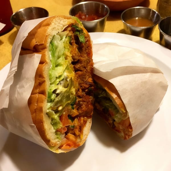 A Mexican torta sandwich, cut in half, revealing meat, lettuce, and sauce, on a white plate.